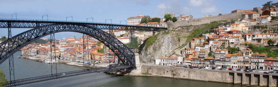 A panoramic view of Porto, Portugal, featuring the iconic Luís I Bridge, colorful buildings, and a historic castle perched on a hill. Porto Tours, Travel Escapes Wine Tours and Cruises