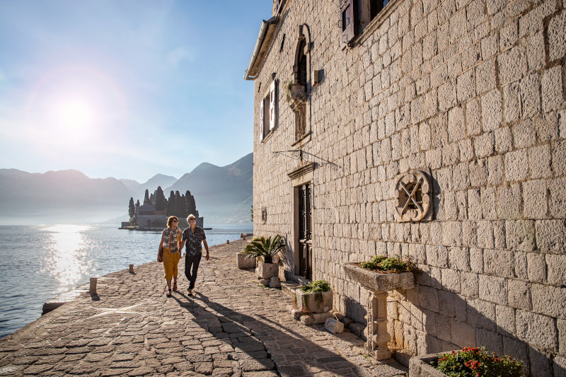 A couple strolls along a cobblestone promenade in Perast, Montenegro, with a view of the iconic Sveti Đorđe Island and the Bay of Kotor in the distance. Travel Escapes Cruises and Tours