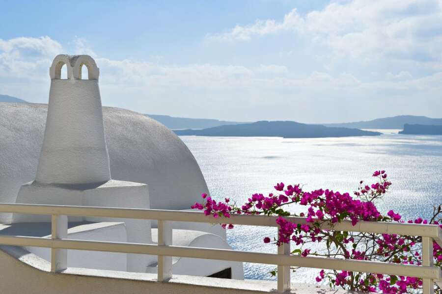 Breathtaking view of the Santorini caldera, featuring a whitewashed building with a distinctive chimney, vibrant bougainvillea, and the sparkling Aegean Sea in the background. Travel Escapes Cruise and Tours