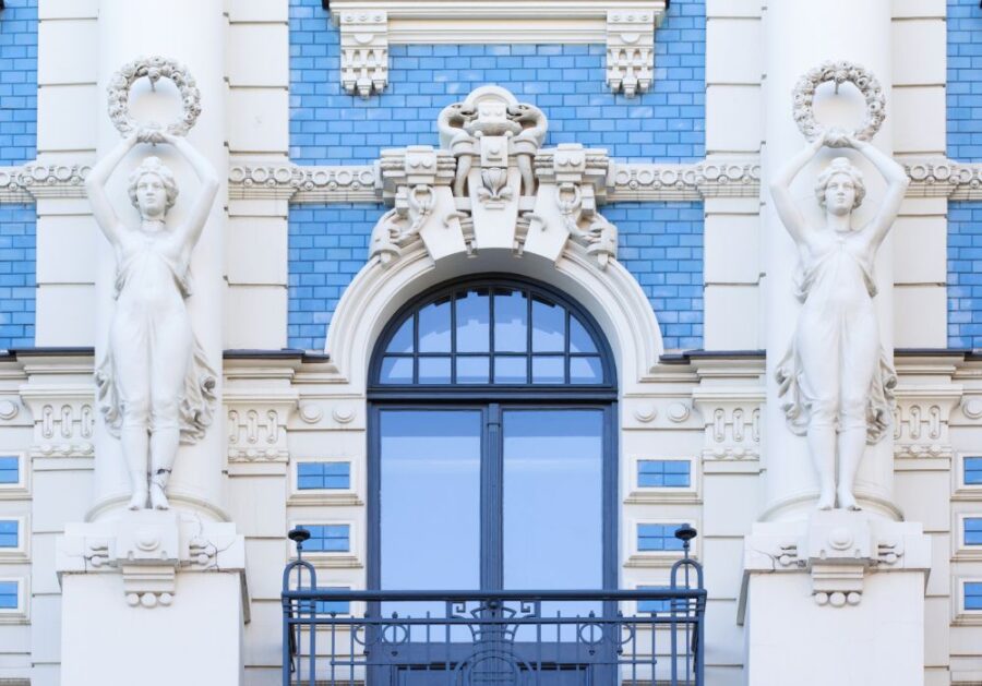 Detailed Art Nouveau facade in Riga, featuring ornate white carvings, blue tile accents, and a dark arched window with balcony. Architectural beauty.