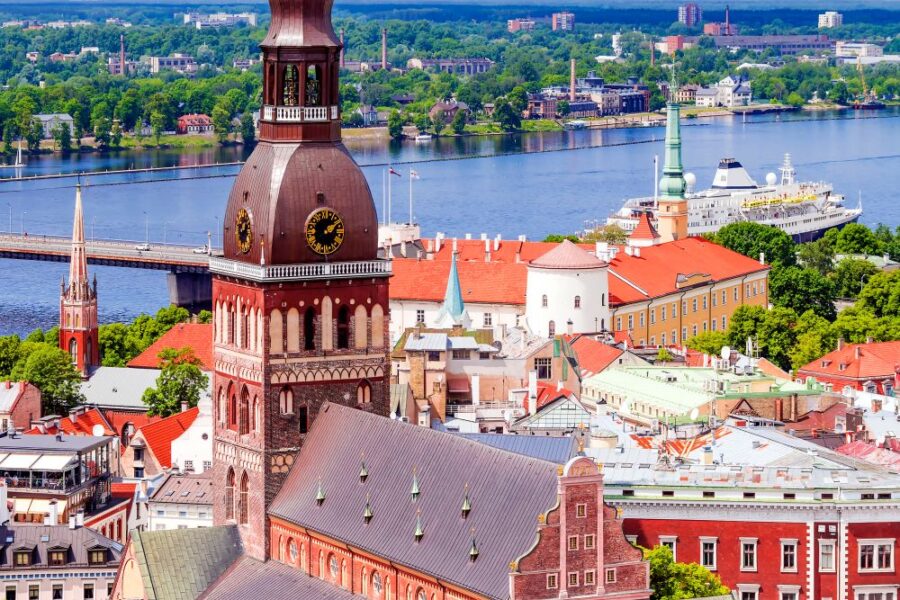 Aerial view of Riga Cathedral, showing its distinctive spire and red-tiled roofs, overlooking the city and river. A panorama of Riga's historic skyline.
