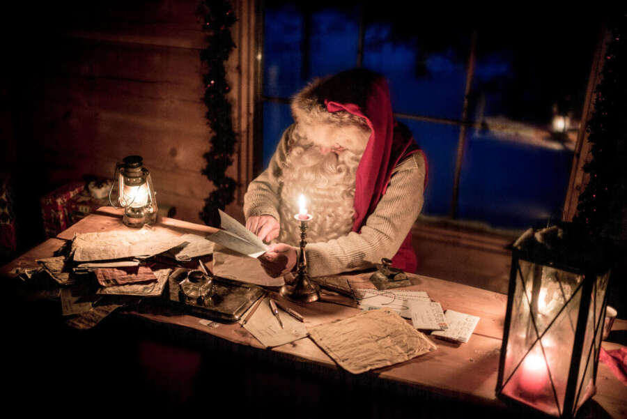 Santa sitting in an office checking his lists in a candle lit room.