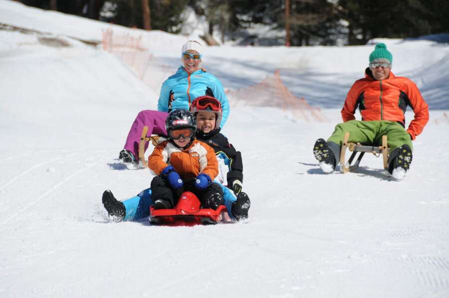 A family enjoys sledding down a snowy hill, smiling and dressed in colourful winter gear. Travel escapes.