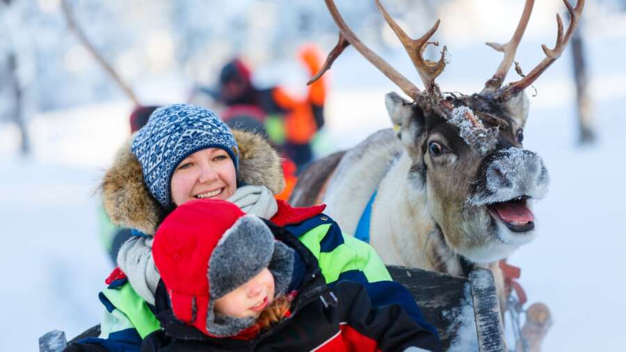 Smiling woman and child enjoy a reindeer sled ride through snowy Lapland, with a reindeer in focus.