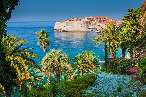 View of Dubrovnik, Croatia, showcasing the iconic Old Town surrounded by fortified walls, with lush palm trees and the Adriatic Sea in the foreground. Travel Escapes Cruises and Tours 