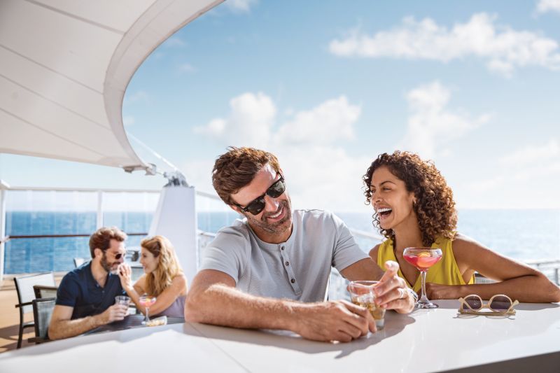 A couple enjoys cocktails on the deck of a Mediterranean cruise, soaking up the sun with the ocean stretching behind them.