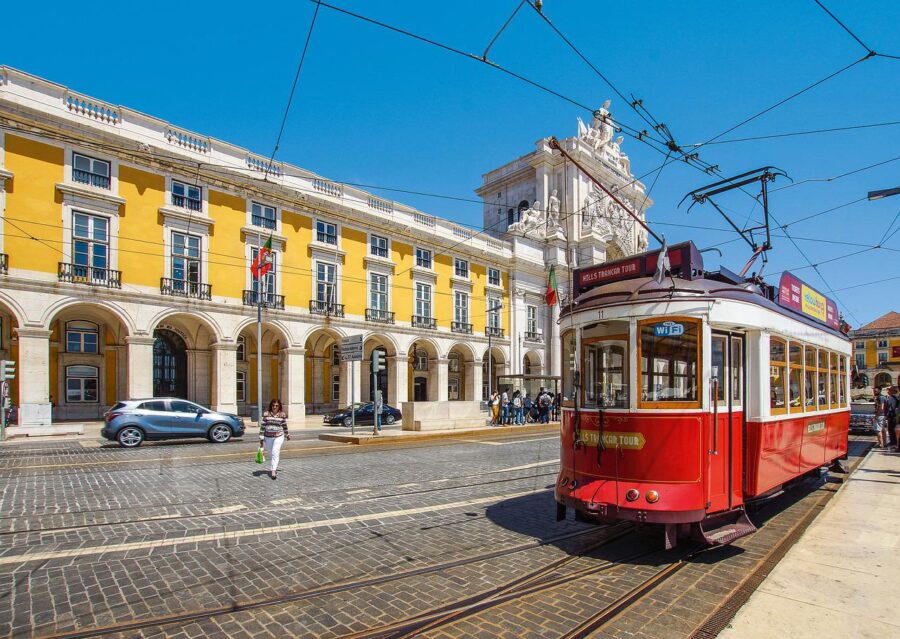 A classic red tram passing by a historic building in Lisbon, Portugal. The tram is a popular mode of transportation and a major tourist attraction in the city. Travel Escapes Escorted Tours & Cruises