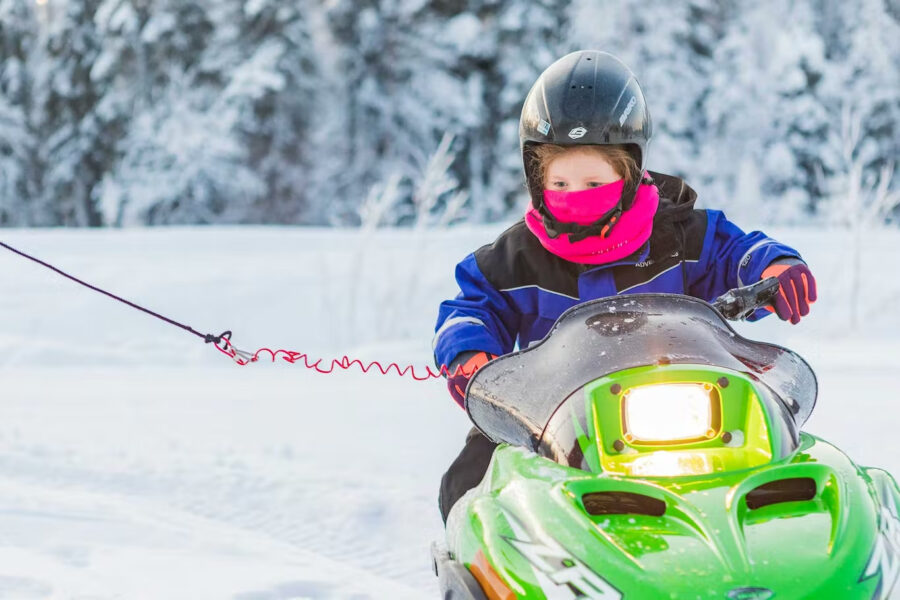 Young child riding a bright green snowmobile, wearing winter gear and a helmet, in a snowy forest.