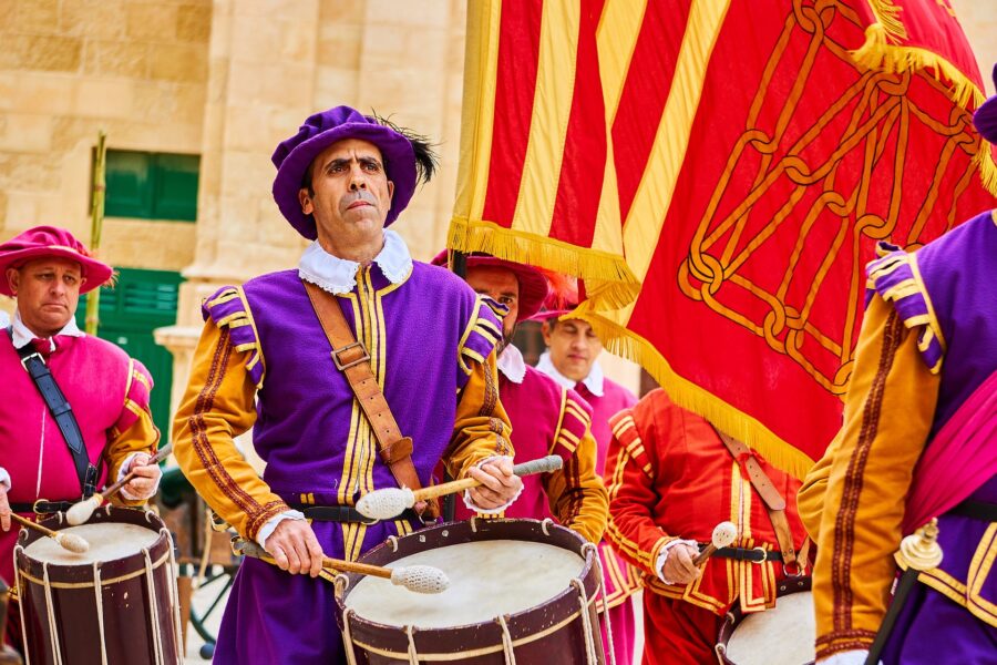 A drummer in a vibrant purple and gold uniform plays a large drum in Malta. Part of a historical or cultural performance, with colourful flags in the background.