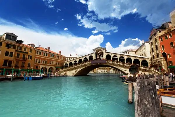 A picturesque view of Venice, Italy, featuring the iconic Rialto Bridge spanning the Grand Canal, with colorful buildings lining the waterfront and gondolas navigating the bustling waterway. Travel Escapes Cruises and Tours