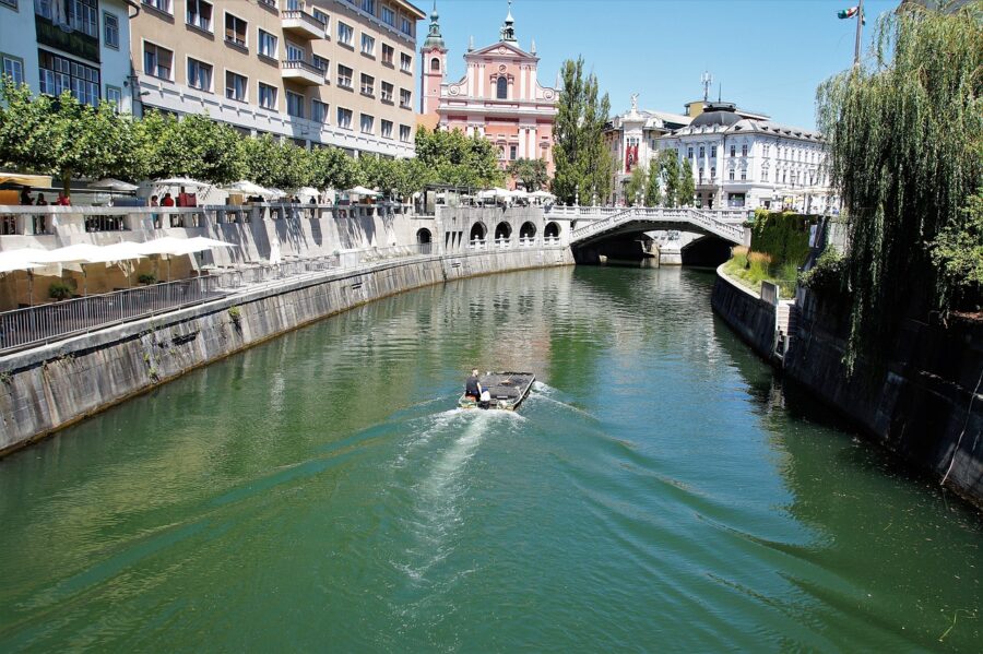 View of the Ljubljanica River in Ljubljana, Slovenia, with a boat gliding along the green water. The iconic Triple Bridge and the pink Franciscan Church of the Annunciation are seen in the background, surrounded by historic buildings and riverside cafes. Travel Escapes Tours and Cruises