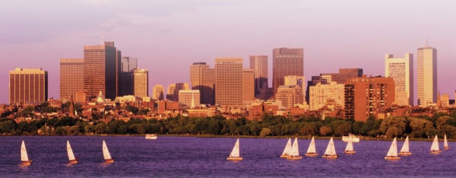 Sailboats on a shimmering river with a modern city skyline bathed in warm evening light.