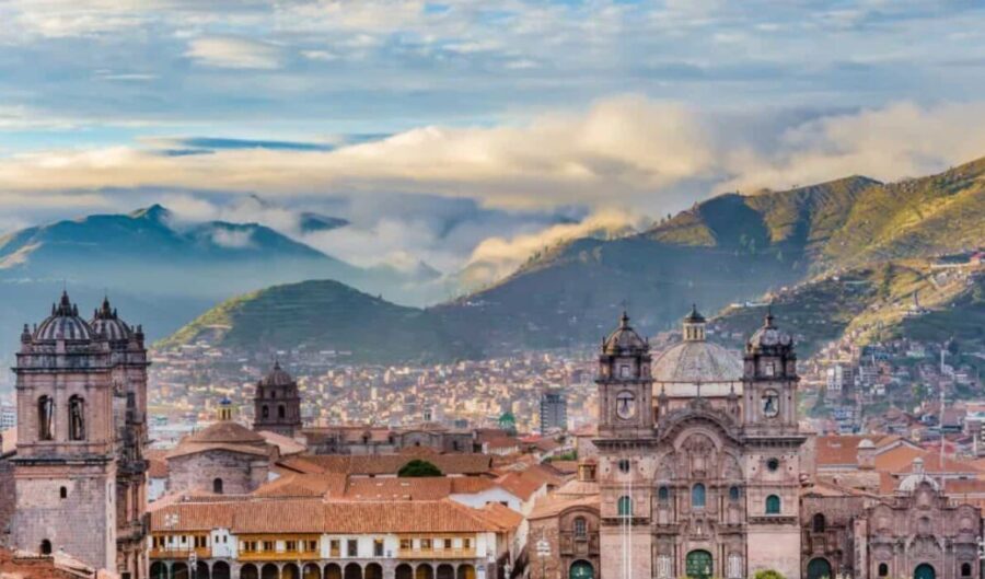 Cusco cityscape, Peru. Colonial architecture, red tile roofs, mountains in background, cloudy sky. Blurry foreground.