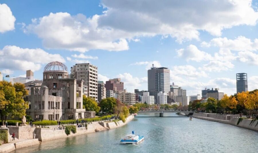 Hiroshima cityscape, Japan. River scene with modern buildings, A-Bomb Dome visible in distance. Blue sky, some clouds.