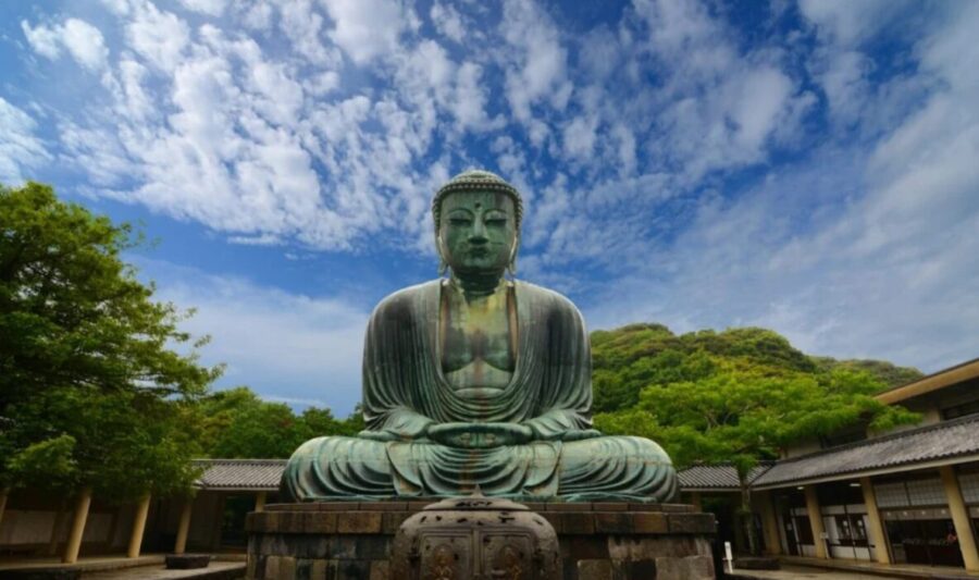 Giant bronze Buddha statue (Great Buddha of Kamakura) seated outdoors, trees behind, blue sky. Japan.