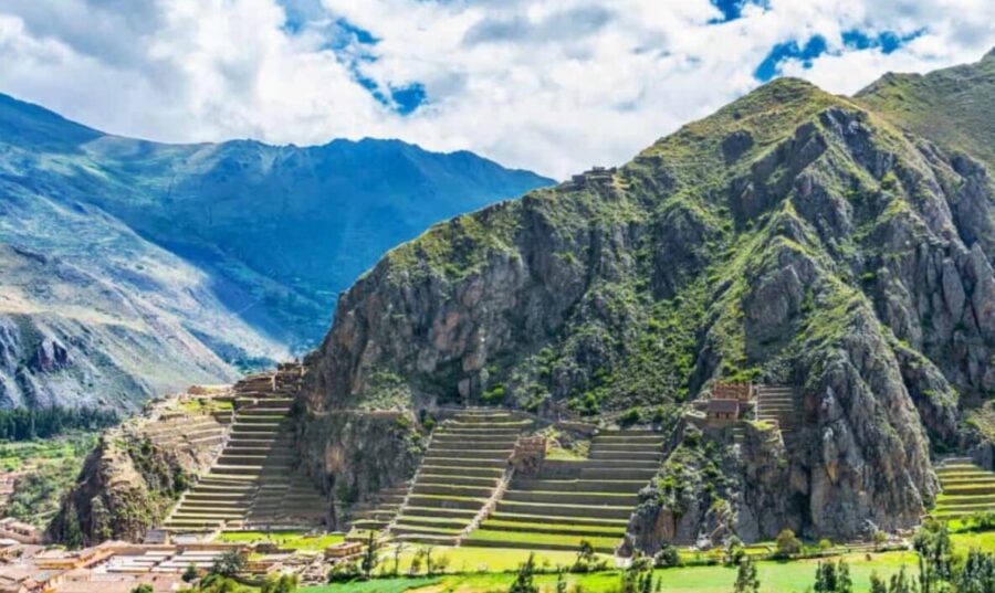 Ollantaytambo, Peru. Inca terraces and ruins on steep hillside, green valley below, mountains in background, cloudy sky.