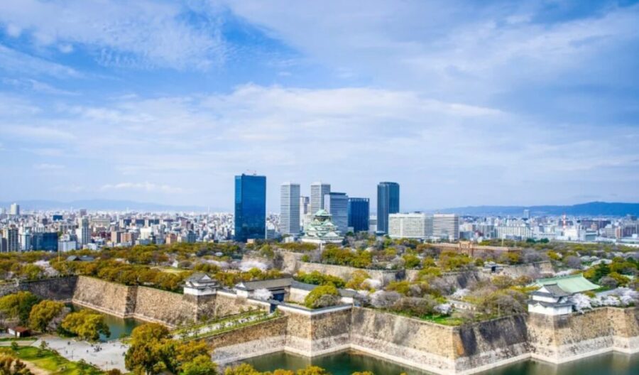 Osaka cityscape, Japan. Osaka Castle walls and moat in foreground, modern skyline with skyscrapers behind. Blue sky.