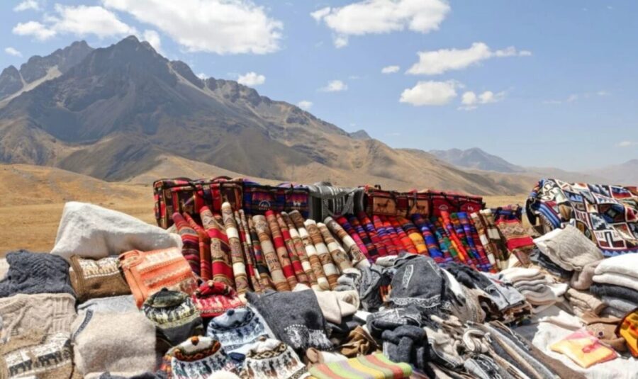 Mountain market scene. Colourful textiles and goods displayed on ground, vendor stalls, hills and sky in background.