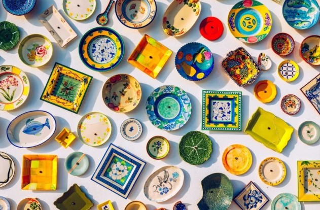 A colourful array of Portuguese plates and tiles displayed on a white surface. Various shapes, sizes, and intricate patterns in blues, yellows, greens, and reds.