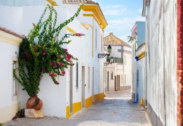 A charming Algarve street with white-washed buildings, yellow trim, and vibrant pink flowers spilling from a pot. Sunny, narrow lane evokes a relaxed holiday vibe.