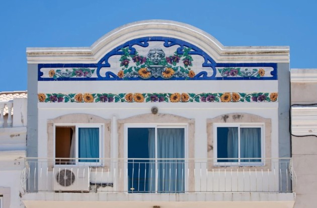 Algarve house with white facade, blue decorative tiles above windows and a curved arch. Building features sliding glass doors and a small balcony. Bright, sunny day.