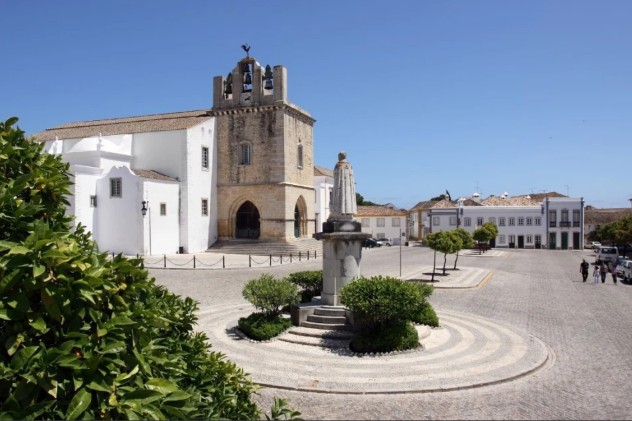 Faro Square with a stone monument at its centre. A large, light-coloured building with a bell tower stands in the background. Green trees and a clear blue sky.