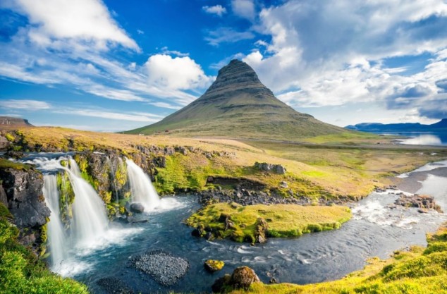 Marvel at the majestic Kirkjufell mountain in Iceland, a striking peak rising dramatically above the surrounding landscape. Cascading waterfalls flow gently in the foreground, creating a picturesque scene under a partly cloudy blue sky. This iconic landmark on the Snæfellsnes peninsula is a photographer's dream and a symbol of Iceland's breathtaking natural beauty.
