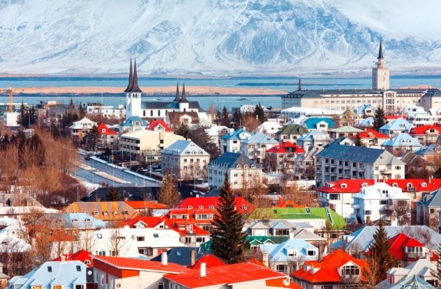 A colourful panoramic view of Reykjavik, Iceland's capital city, showcasing its charming architecture with brightly coloured rooftops and the prominent spire of Hallgrímskirkja church. In the background, majestic snow-capped mountains rise against a blue sky and the waters of the coastline. This vibrant urban landscape offers a glimpse into Icelandic city life and its stunning natural surroundings.