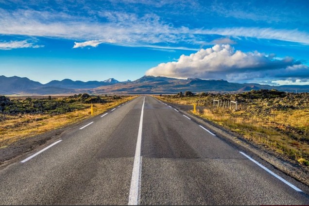 An open road stretches into the distance under a bright blue sky dotted with fluffy white clouds, typical of Iceland's Golden Circle route. The stark yet beautiful landscape features low-lying vegetation on either side, leading towards distant mountains on the horizon. This image captures the sense of freedom and adventure of exploring Iceland's iconic scenic drive.