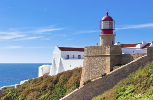 Sagres lighthouse with red dome against a clear blue sky. Stone walls and white buildings visible. Ocean in the background. Sunny coastal scene.