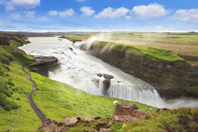 The majestic Gullfoss waterfall in Iceland showcases its impressive two-tiered cascade as glacial water thunders down into a rugged canyon. Lush green vegetation surrounds the falls under a vibrant blue sky with scattered clouds. This powerful and beautiful natural wonder is a highlight of Iceland's Golden Circle and a testament to the country's dramatic landscapes.