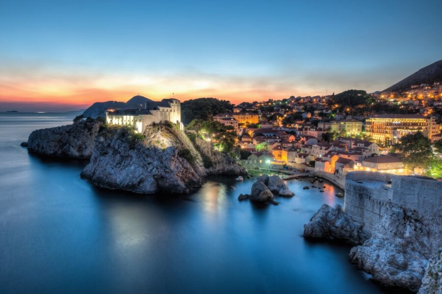 The ancient walls of Dubrovnik glow softly at dusk, overlooking the still, dark blue Adriatic Sea. City lights twinkle along the coastline as the sky blends hues of orange and blue. Discover the beauty of this historic Croatian gem.