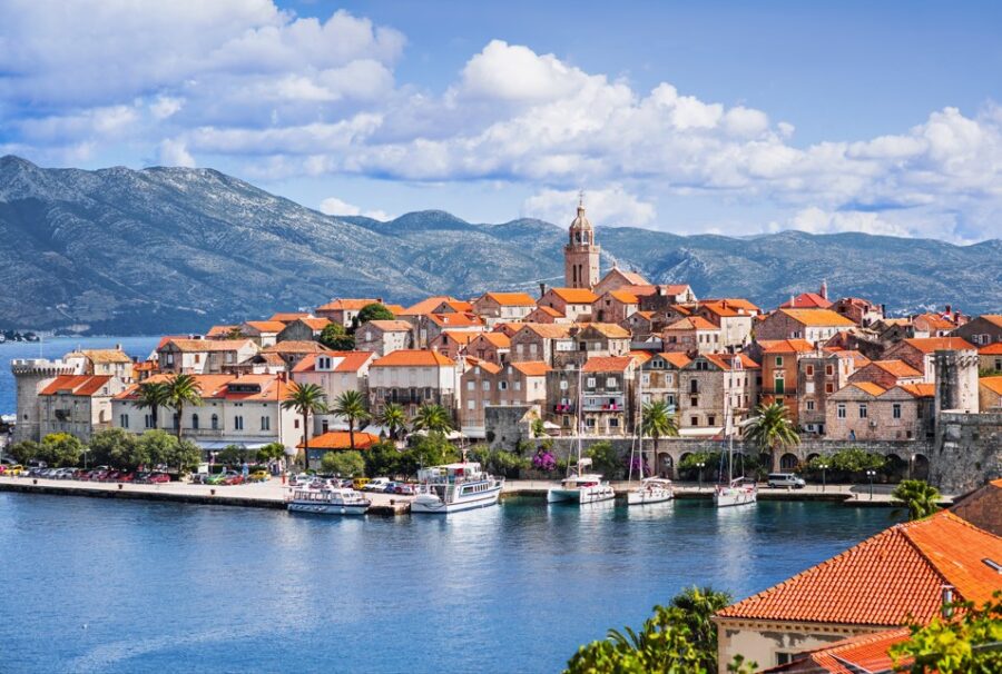 A picturesque view of Korčula town, with its terracotta rooftops clustered within medieval walls beside the clear blue Adriatic Sea. A prominent cathedral tower rises above the charming Croatian settlement under a partly cloudy sky. Explore the beauty of the Dalmatian Islands.