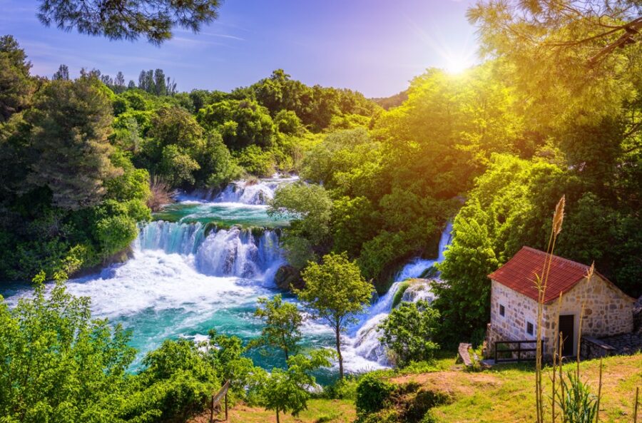 Stunning turquoise waterfalls cascade through lush green vegetation under a bright, sunny sky in Krka National Park, Croatia. Sunlight glints on the flowing water, creating a vibrant and inviting scene. Discover the natural wonders of the Dalmatian Coast.