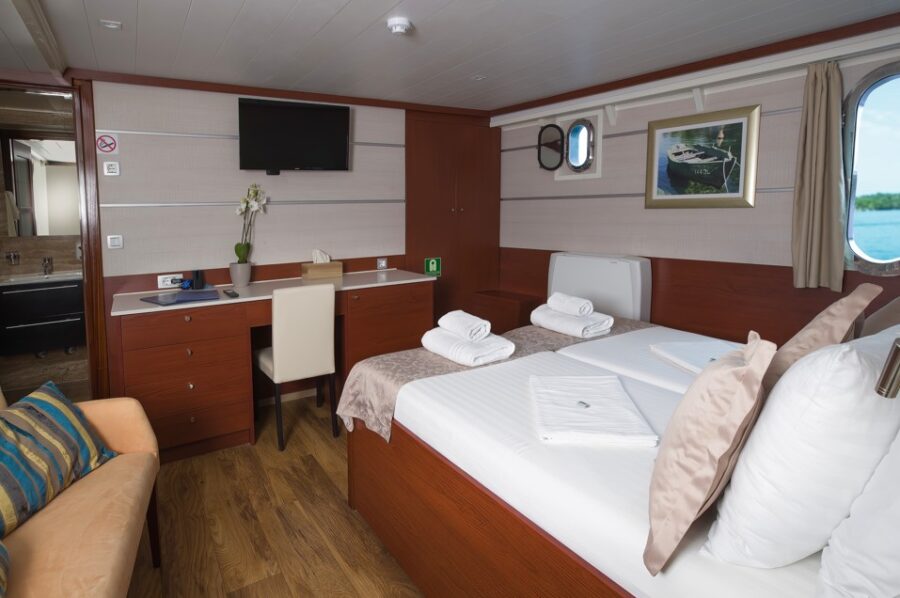 A cosy cabin on the Princess Eleganza featuring a double bed with fresh white towels, a wooden bedside table, a desk area with a chair, and a mounted TV. A porthole window lets in natural light. Enjoy comfortable cruising on the Adriatic.