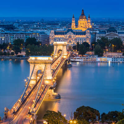 Iconic Budapest Chain Bridge beautifully illuminated at night over the Danube, with St. Stephen's Basilica glowing in the background.