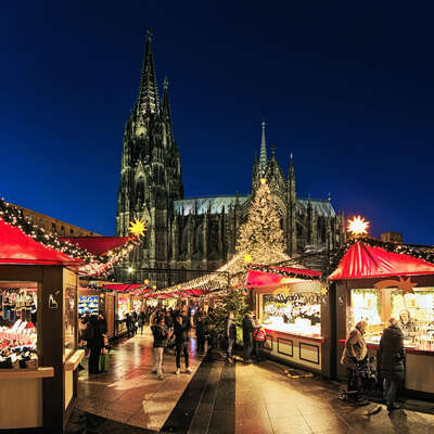 Bustling German Christmas market at night, traditional wooden stalls glowing with lights, set against the majestic Cologne Cathedral.