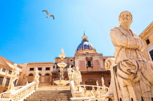 Explore Palermo's stunning Fontana Pretoria! Ornate statues and a grand domed building gleam under a brilliant blue sky, with a soaring seagull. Iconic Sicilian beauty.