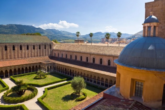 Discover the peaceful Monreale Cathedral Cloister in Sicily. Serene courtyard gardens, elegant arcaded walkways, and majestic mountains under a clear blue sky. A stunning historic gem.