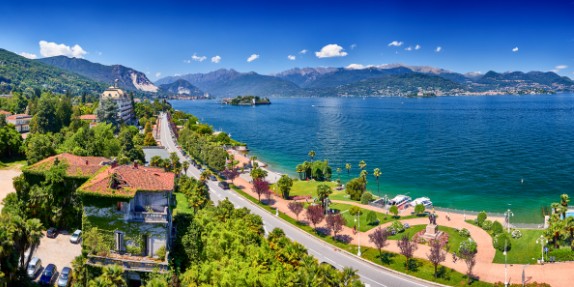A beautiful view of Lake Maggiore in Italy, with a tree-lined promenade, deep blue water, and the Borromean Islands and mountains in the distance.