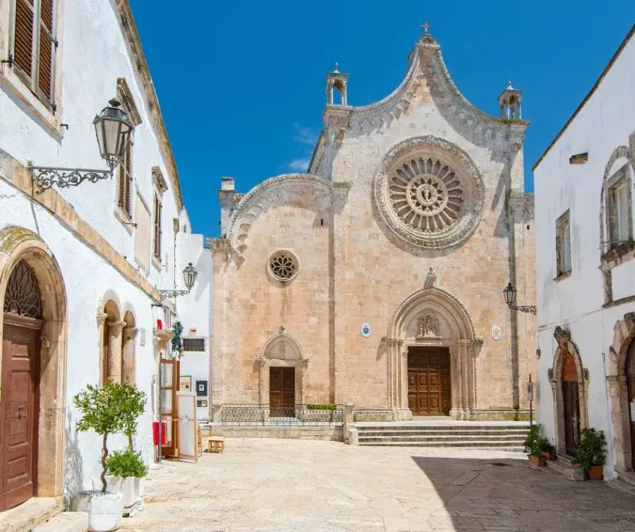 The facade of the 15th-century Ostuni Cathedral in Puglia, Italy, showing its intricate rose window, set against a bright blue sky.